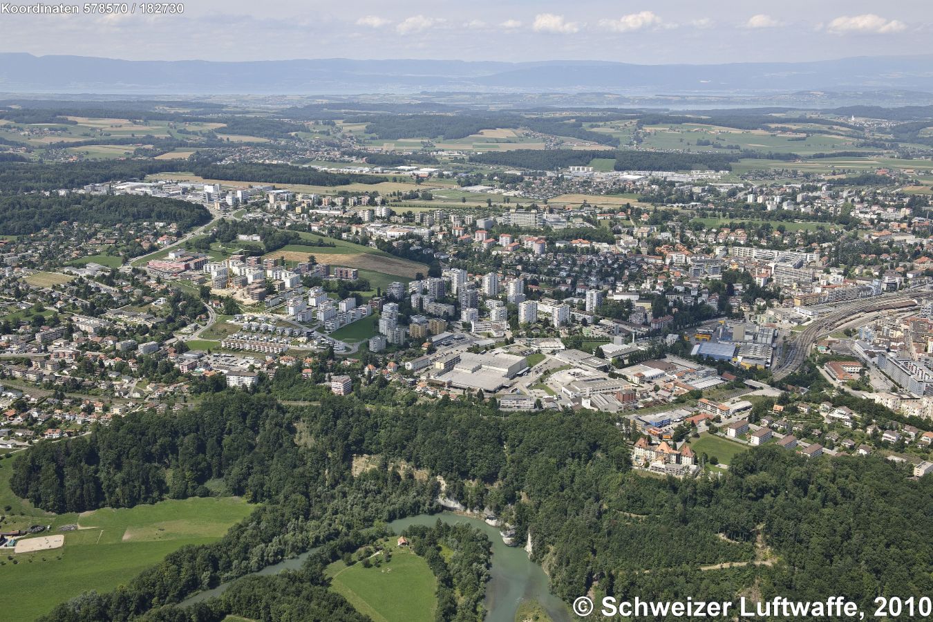 Fribourg Übersicht. Rechts Bildmitte: Bahnhof SBB. Im Vordergrund Prallhang der Sarine ('Creux -du - Loup), darüber im 'Bois des Morts' die Villa Beausite (Position 2'577'916.04, 1'182'351.35). - Im Bildhintergrund Neuenburger- und Murtensee. Rechts: Villars sur Glâne. Im Mittelgrund: Givisiez.